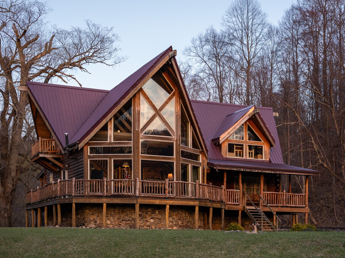 A spacious log cabin is showcased, featuring a prominent wooden exterior with large windows that offer views of the surroundings. The steeply sloped roof adds character, while the expansive deck invites relaxation. Natural landscaping surrounds the cabin, enhancing its connection to the outdoors.