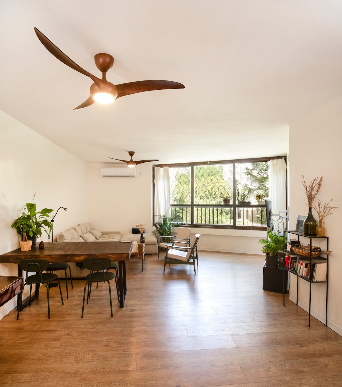 A light-filled living space features modern furnishings, including a wooden dining table surrounded by black chairs. A comfortable sofa is positioned nearby, with greenery and decorative items on a shelf. Large windows invite natural light and provide views of the surrounding area.