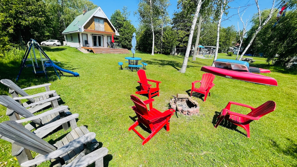 A spacious outdoor area features a firepit surrounded by red Adirondack chairs, inviting relaxation. A blue picnic table sits nearby, with a children's swing set and two kayaks visible in the grass. The cottage's wooden deck and lush trees create a serene backdrop.