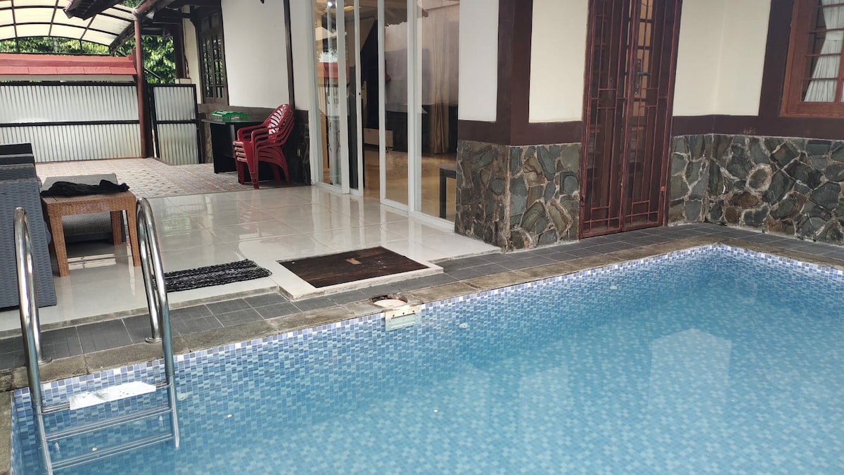 A private pool is framed by smooth stone tiles, reflecting light with a vibrant blue hue. Steps lead into the water, while a nearby seating area includes a table and chairs. Glass doors open to create an inviting connection to indoor spaces.