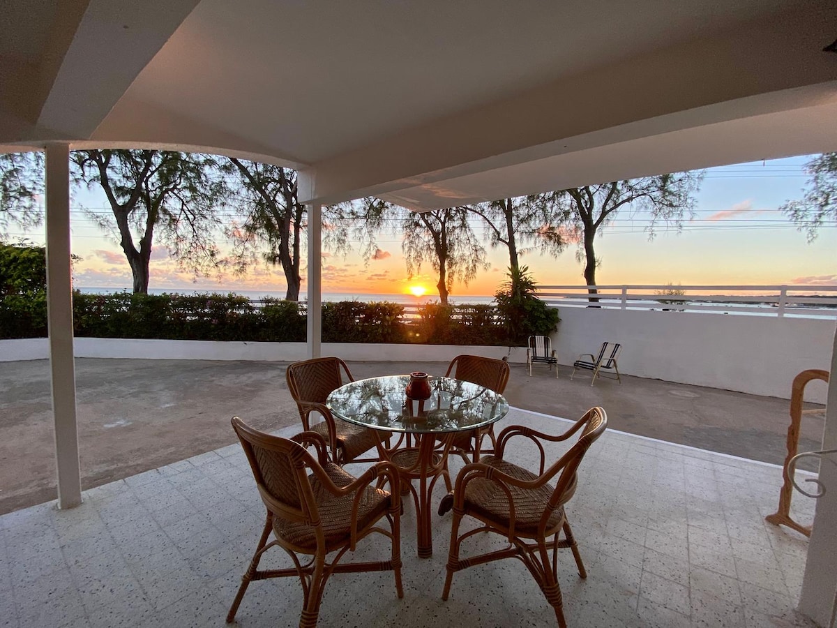 An outdoor patio area features a glass-topped dining table surrounded by four wicker chairs. The space is framed by trees, with a sunset view visible through the open area. Soft lighting highlights the peaceful ambiance of the setting.
