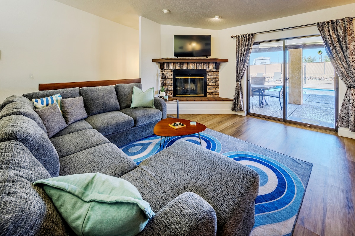 A living area is showcased with a large sectional sofa arranged around a wooden coffee table. A flat-screen TV is mounted above a stone fireplace. Light drapes frame the sliding glass doors, which provide a view of the outdoor patio and pool area, enhancing the spacious feel.