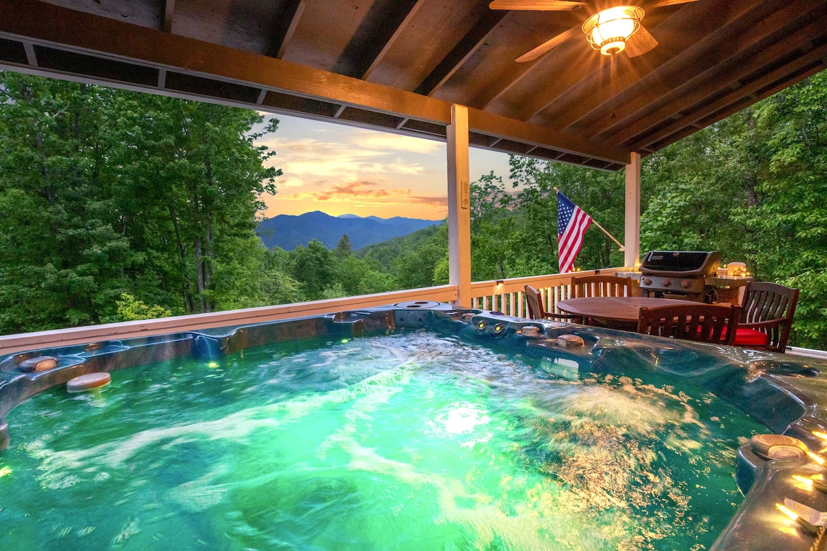 A hot tub filled with bubbling water is situated under a covered porch, surrounded by trees. Scenic views of the mountains are visible in the distance, while a gas grill and a seating area are positioned nearby. An American flag adds a patriotic touch.