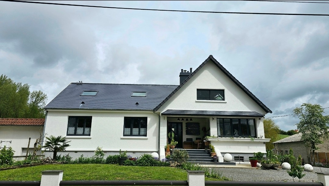 The exterior of a two-story house is presented, featuring a predominantly white facade with a dark slate roof. Large windows provide natural light, surrounded by well-maintained greenery and decorative stones. The entrance is highlighted by a set of steps leading to a welcoming front door.