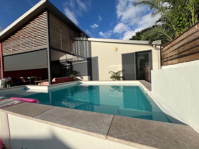 A modern swimming pool is surrounded by a clean stone edge, reflecting the clear blue sky. The property features large windows and a partially shaded outdoor area with seating. Lush greenery adds to the serene environment, inviting relaxation.