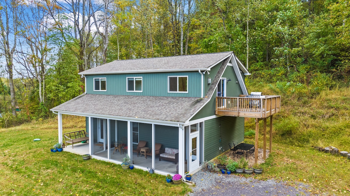 The cottage features a two-story structure with a welcoming porch and screened-in area. A wooden deck extends from the upper level, providing access to outdoor seating. Surrounding trees offer a natural backdrop, and the grassy area includes potted plants for added charm.