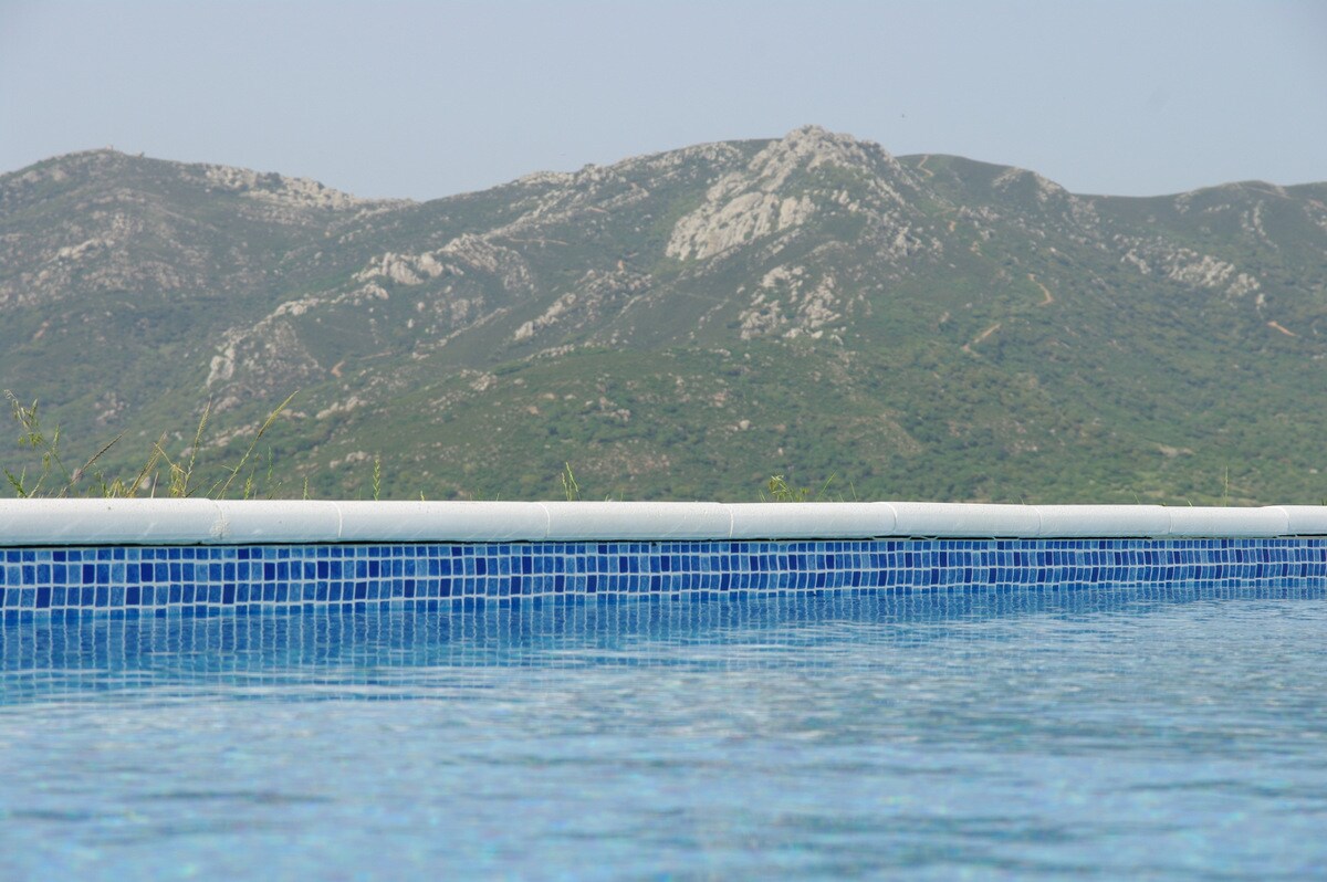 A close-up view of a swimming pool's edge reflects the blue tiles, with a backdrop of green mountains under a clear sky. The pool's surface glistens under sunlight, offering a serene connection to the surrounding natural landscape.