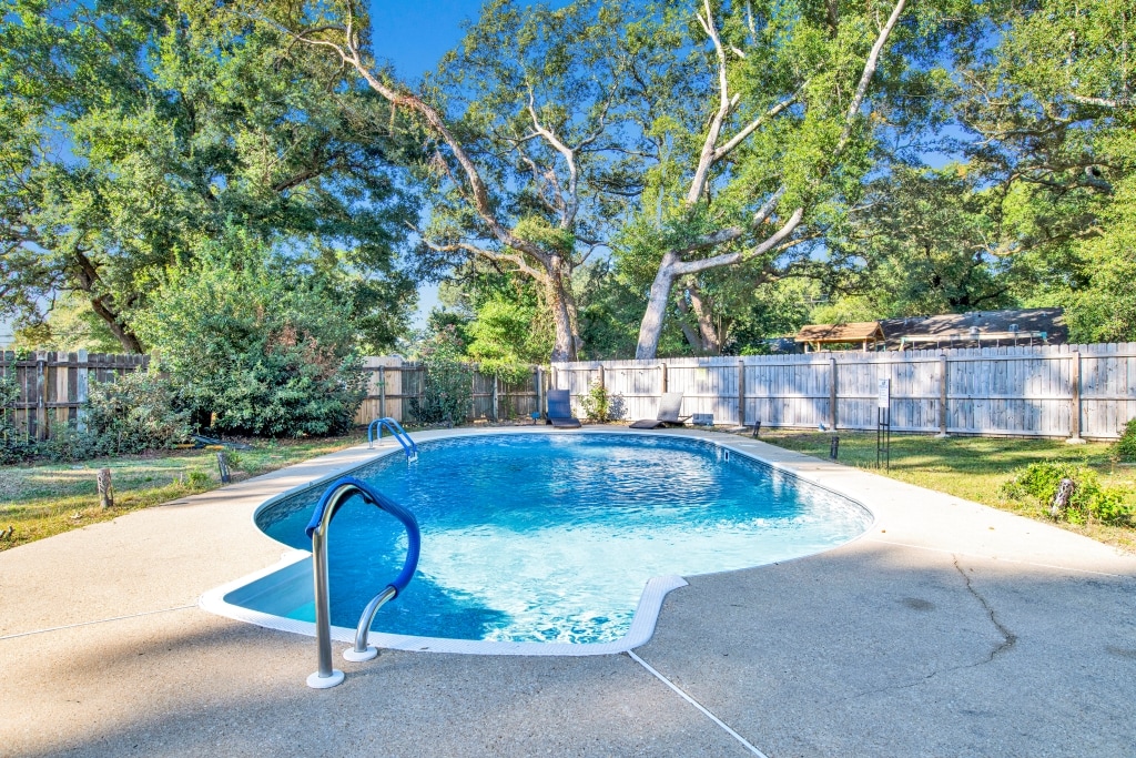 A clear, inviting saltwater pool is surrounded by a spacious patio, featuring a gentle curve and easy access steps. Lush greenery and tall trees frame the area, providing a serene outdoor escape. A wooden fence enhances privacy, while ample sunlight brightens the scene.