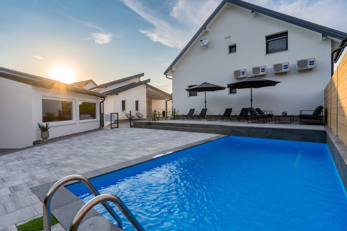A swimming pool is situated in the foreground, featuring clear blue water reflecting the evening sun. A spacious patio area is lined with sun loungers and umbrellas, while the surrounding buildings include large windows and air conditioning units, providing a modern aesthetic.