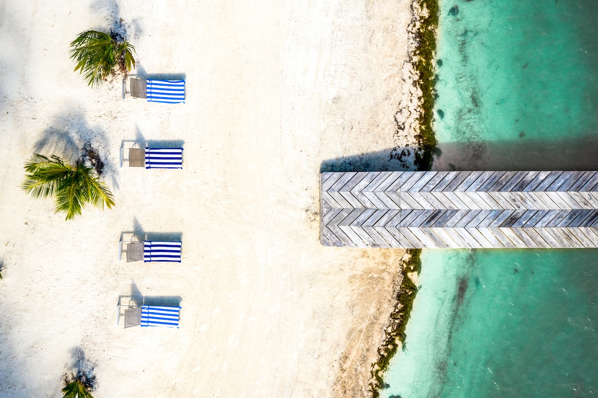 Aerial view of a sandy beach featuring four striped lounge chairs arranged neatly beside a private pier. The pier extends into the turquoise waters, bordered by lush palm trees and the gentle waves of the Caribbean Sea.