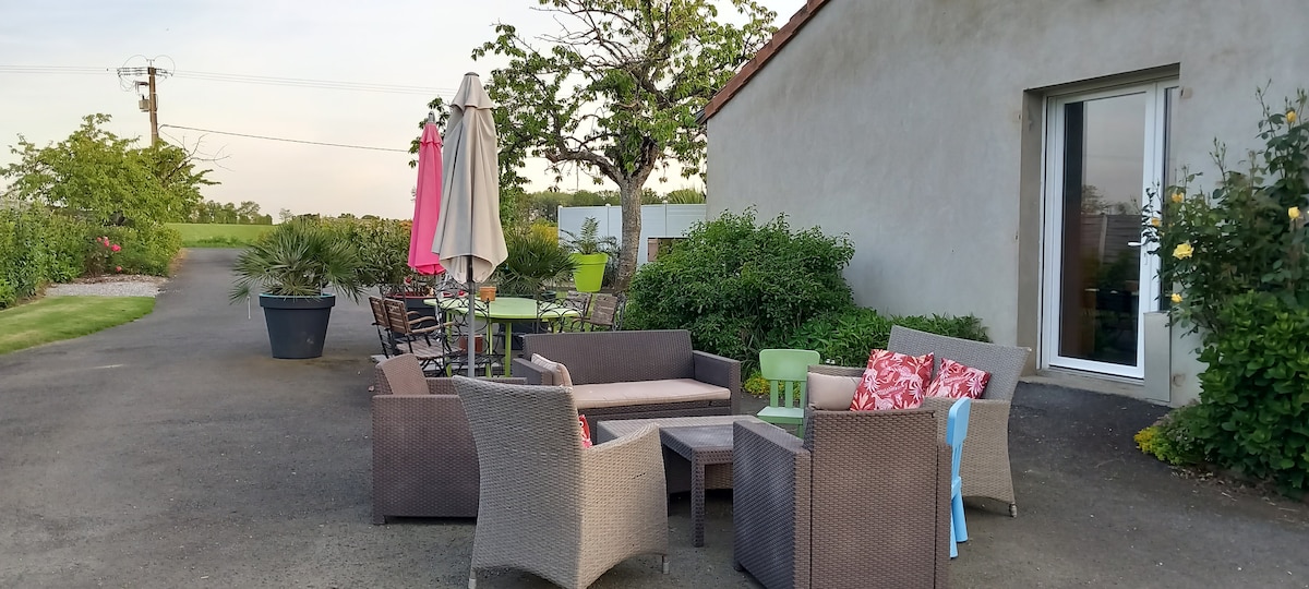 An outdoor seating area features a collection of comfortable wicker furniture, including a sofa and armchairs arranged around a low table. A shade umbrella and decorative plants are present, contributing to a relaxed and inviting ambiance. The space is bordered by greenery and nearby pathways.