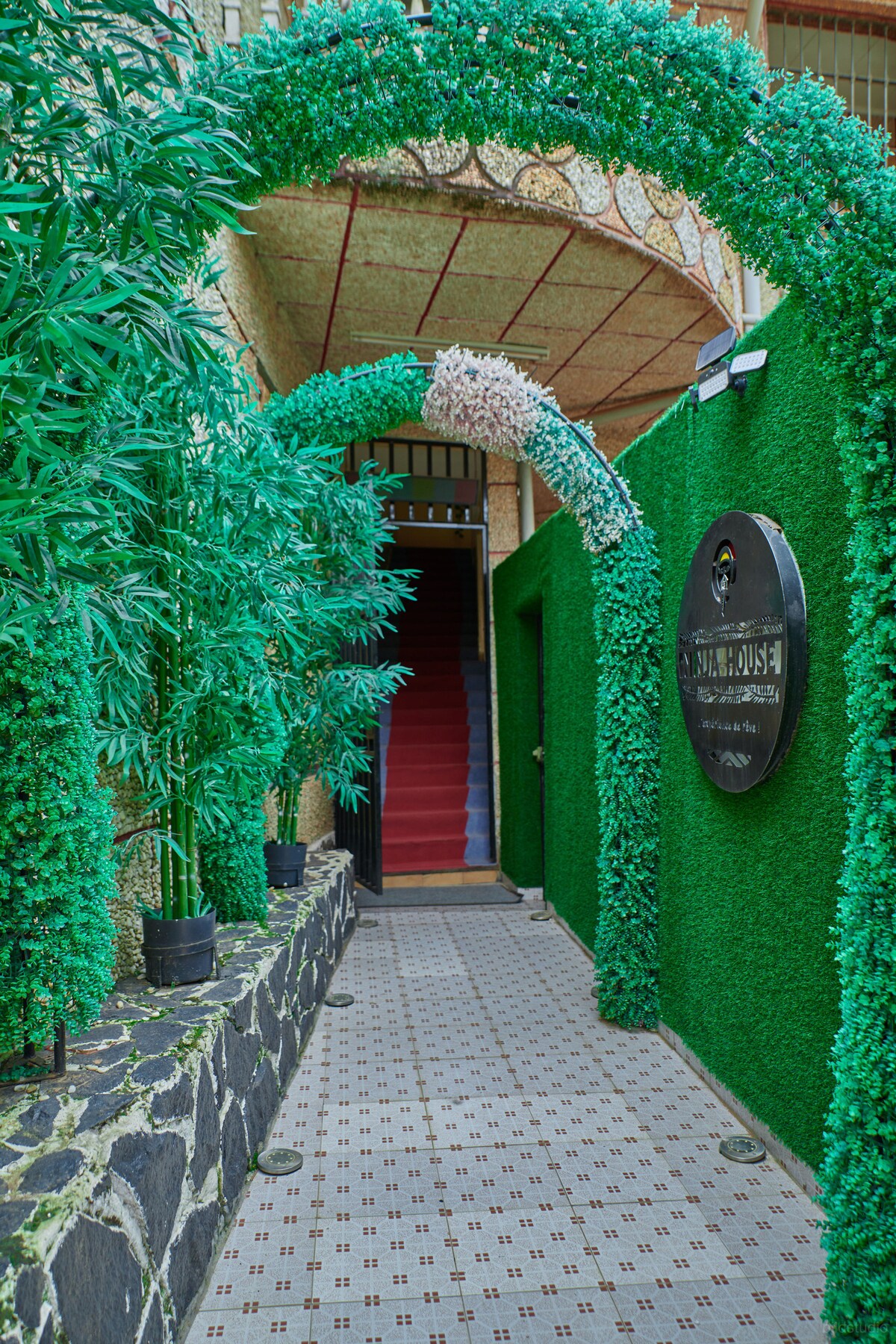 A pathway leads to a staircase surrounded by lush green archways. The entrance showcases a decorative sign, and a textured stone wall complements the vibrant foliage, creating a welcoming approach to the building.