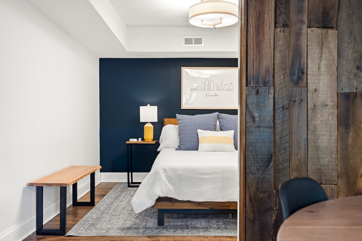 A serene bedroom is highlighted by a dark blue accent wall and warm wooden elements. The spacious bed is adorned with soft pillows and an inviting blanket. A simple bedside lamp and artwork add subtle charm, while natural light fills the room from the doorway.