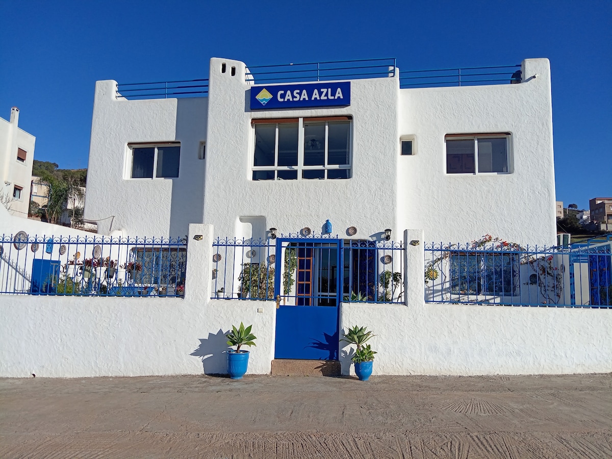 The exterior of Casa Azla is displayed, featuring a white facade and a blue entrance. Decorative plants frame the entrance, and the property is enclosed by a metal fence. A clear blue sky provides a striking backdrop, adding to the inviting atmosphere of the guesthouse.