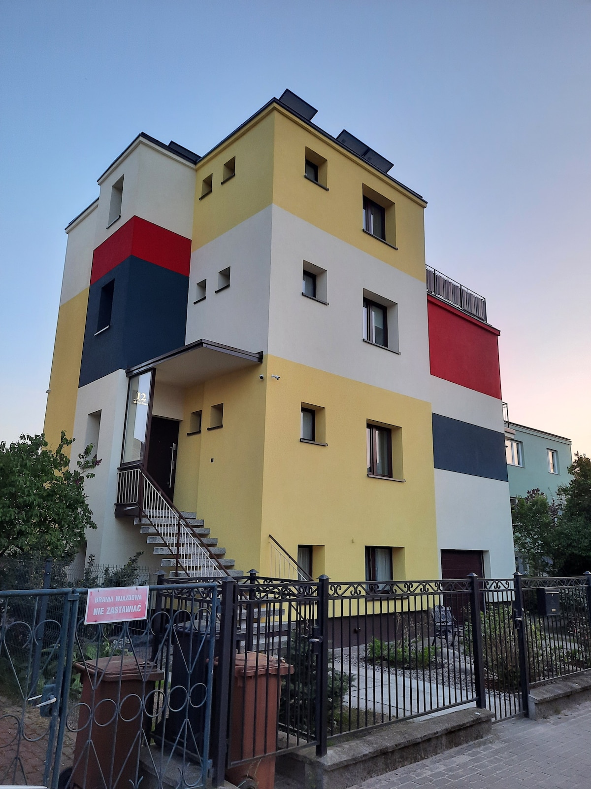 A multi-story, colorful building is shown, featuring a mix of yellow, red, and gray facades. A set of stairs leads to the entrance, framed by a gate and decorative landscaping. The sky reflects warm tones of dusk above the structure.