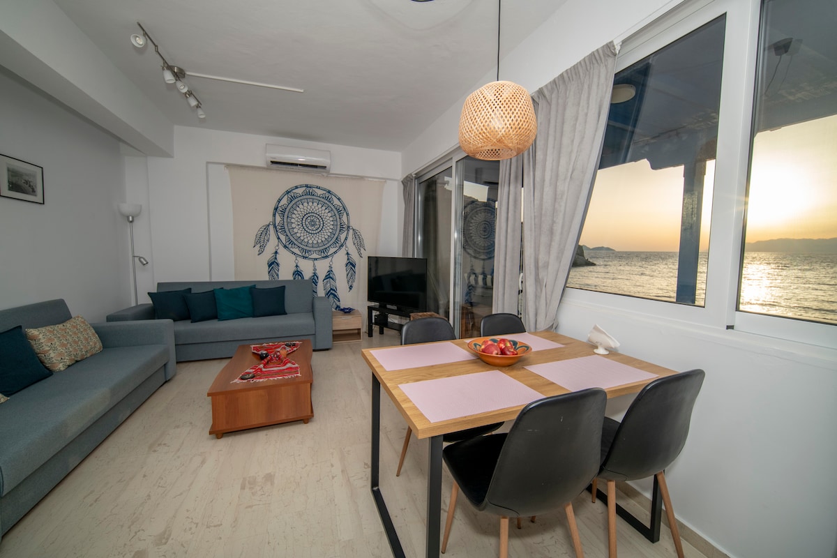 A spacious living area features comfortable seating and a wooden dining table set for meals. Large glass doors reveal an expansive view of the sea and allow natural light to fill the room. Decorative elements include a wall tapestry and indoor plants.