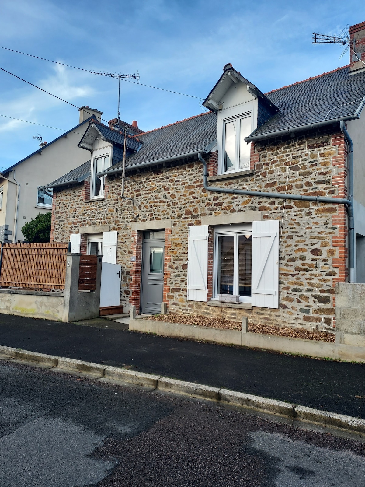 A charming stone house with a historic facade is visible, featuring white shutters and a modern front door. A wooden fence creates a sense of privacy, while the surrounding sidewalk is lined with trees and parked cars, offering a glimpse of the quiet neighborhood.