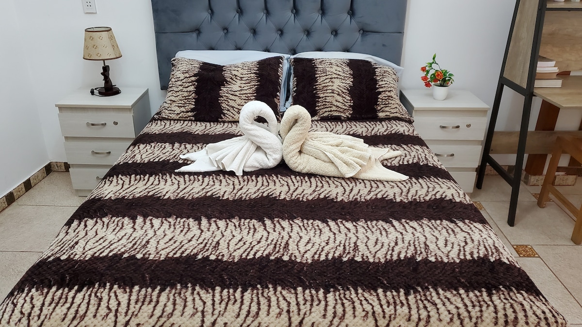 A spacious bed is covered with a decorative blanket featuring a striped pattern in dark and light tones. Two towel swans are elegantly arranged at the center of the bed. Nightstands are positioned on either side, with a small lamp on one and greenery in a pot on the other.