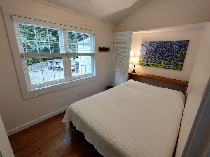 חדר שינה צפוני עם מיטת קווין סייז המשקיף על החצר הצדדית. הציור הוא "אורות גן", שמן על קנבס (2024).