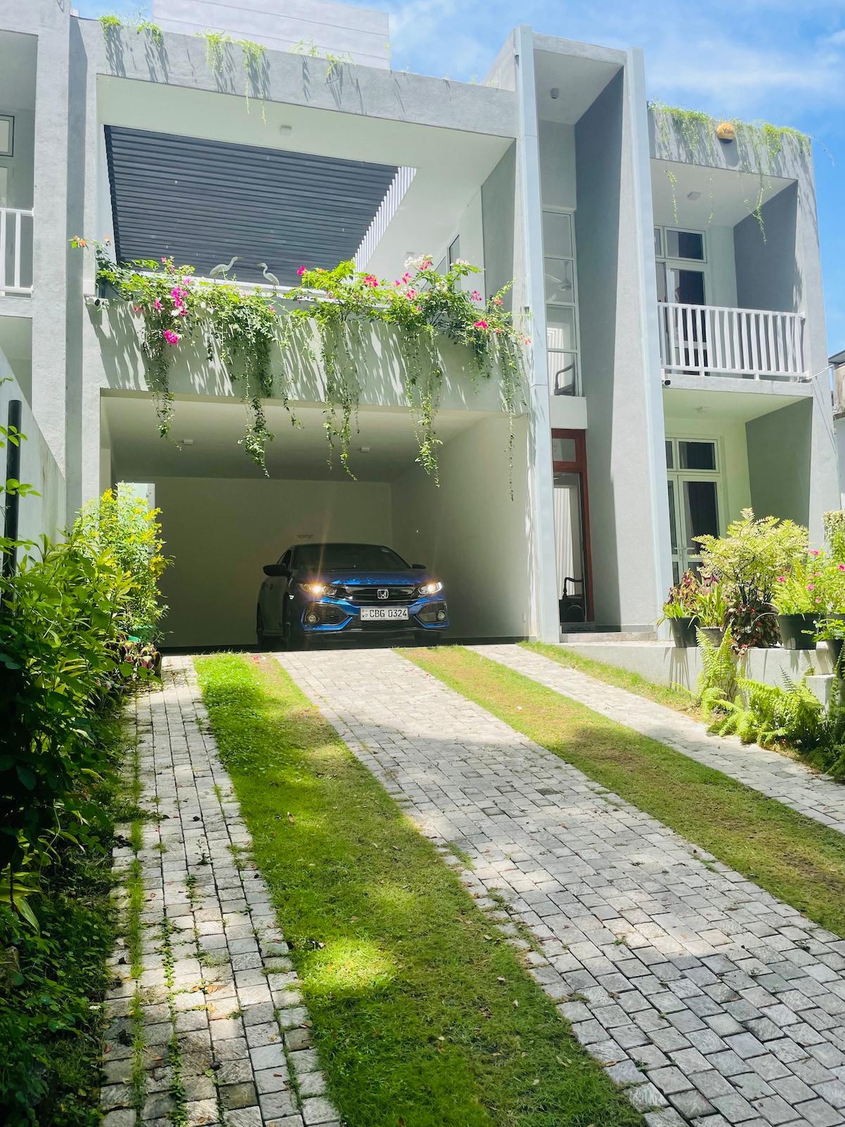 A modern building features a striking facade adorned with vibrant greenery. The driveway, lined with interlocking stones, leads up to a spacious carport filled with natural light. A well-maintained garden area adds to the overall appeal, enhancing the welcoming entrance.