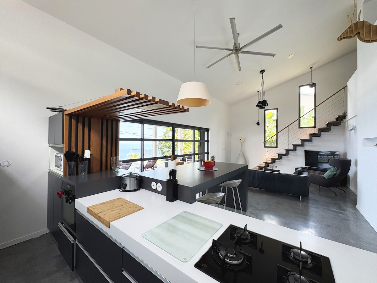 A modern kitchen and living area feature a kitchen island with a stovetop, contrasting cabinetry, and ample natural light. A comfortable couch and stylish decor create a functional space. Large windows offer views of the outdoors, enhancing the open atmosphere.