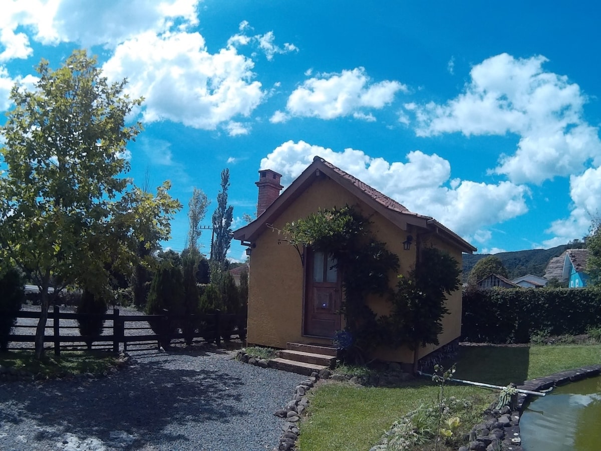 A charming yellow cottage is captured under a bright blue sky with fluffy clouds. The cottage features a stone pathway leading to its entrance, accented by greenery and a climbing vine. Surrounding trees enhance the outdoor space, offering a tranquil setting.