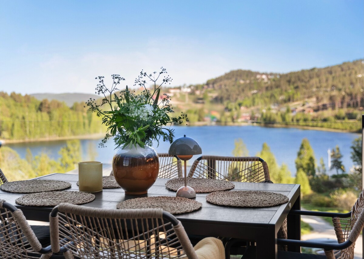 An outdoor dining table is set with natural placemats and a centerpiece featuring fresh flowers. Comfortable wicker chairs surround the table, offering a view of the lake and wooded hills in the background, creating an inviting space for gatherings.