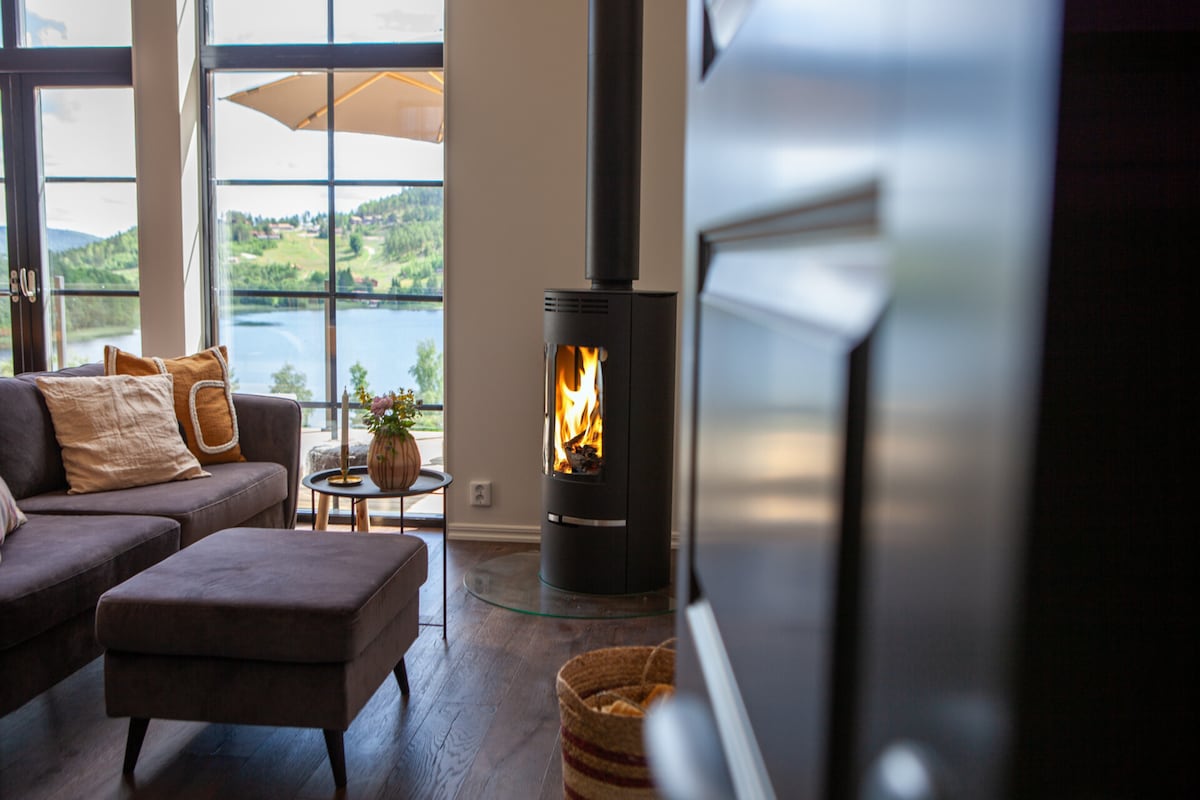 A modern living area features a comfortable sofa and an ottoman, accompanied by a coffee table. A wood-burning stove is located beside large windows that offer views of the lake and surrounding hills. Natural light enhances the ambiance, creating a cozy atmosphere.