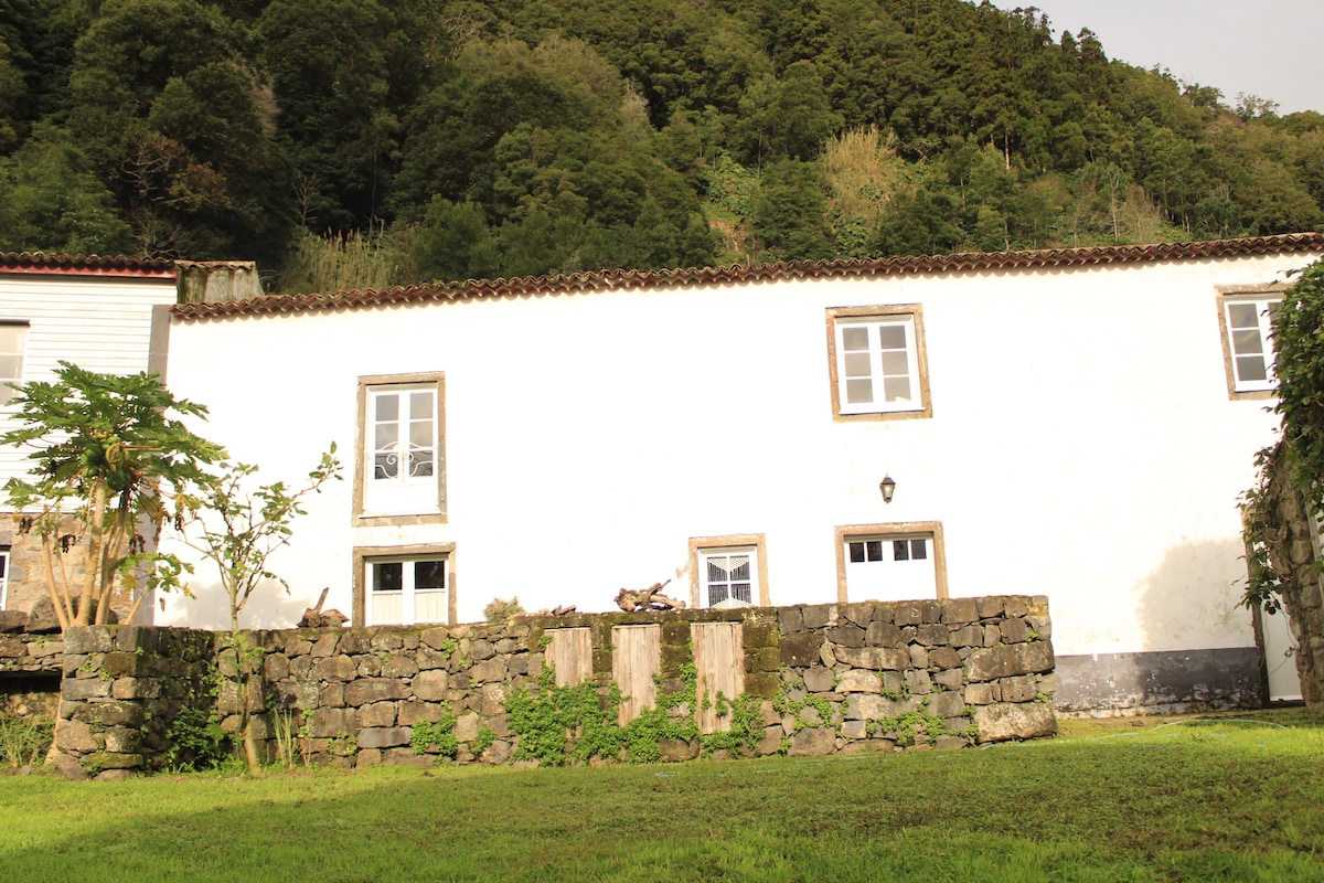 A traditional lava stone house is seen against a backdrop of lush green hills. The structure features white walls and several windows, framed by a stone wall and well-maintained grassy area, creating a harmonious blend with the surrounding nature.