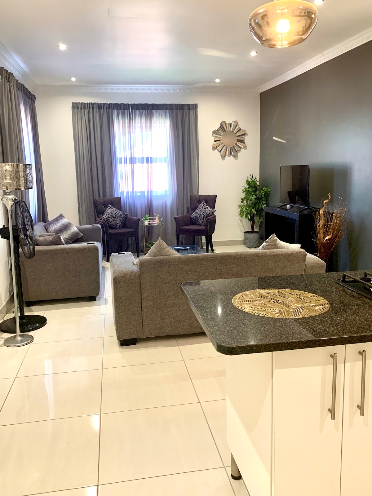 A modern living area is presented, featuring two gray sofas and two accent chairs. Soft lighting illuminates the space, which includes a flat-screen television and a decorative wall mirror. Light-colored curtains softly frame the windows, adding to the inviting ambiance.