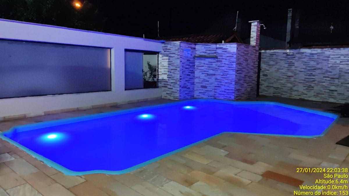 A nighttime view of a swimming pool features illuminated water in soft shades of blue. The pool area is surrounded by stone tile flooring, with a backdrop of textured stone walls. Additional lighting enhances the ambiance of the outdoor space.