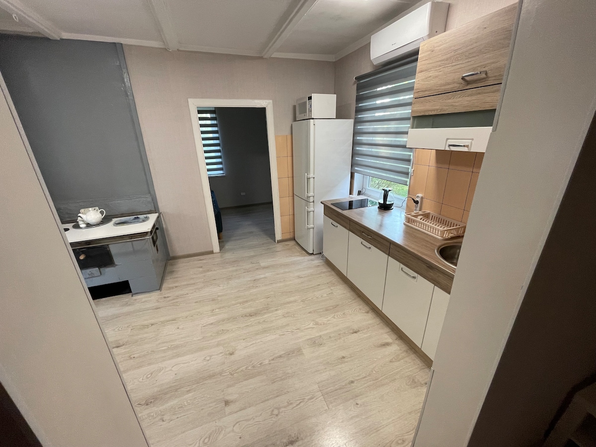 A well-equipped kitchen is shown, featuring a refrigerator and an oven, along with a compact sink and wooden countertop. Light-colored cabinetry contrasts with the gray wall. Natural light enters through window shades, illuminating the wooden flooring throughout the space.
