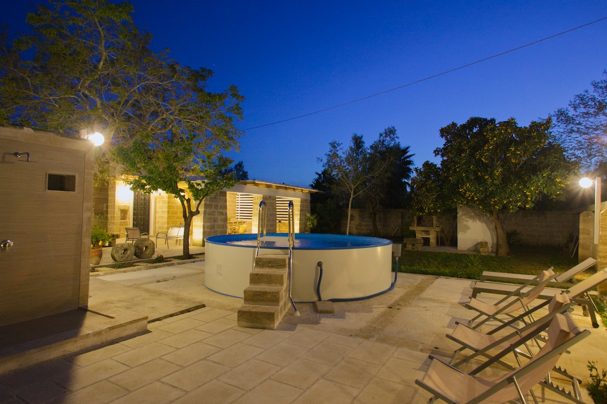 A well-lit pool area is presented under a twilight sky, featuring a circular above-ground pool surrounded by a spacious deck. Comfortable lounge chairs are positioned nearby. An outdoor kitchen and a rustic stone table are also visible, tucked against greenery in the background.