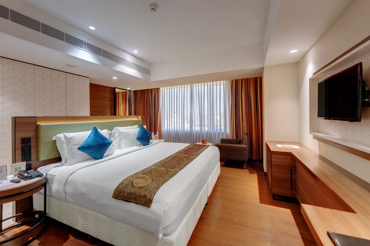 A well-appointed executive room features a spacious bed with decorative throw pillows and an elegant bed runner. Natural light filters through wide curtains. A stylish seating area is present, along with a wall-mounted TV and a wooden desk positioned against the wall.