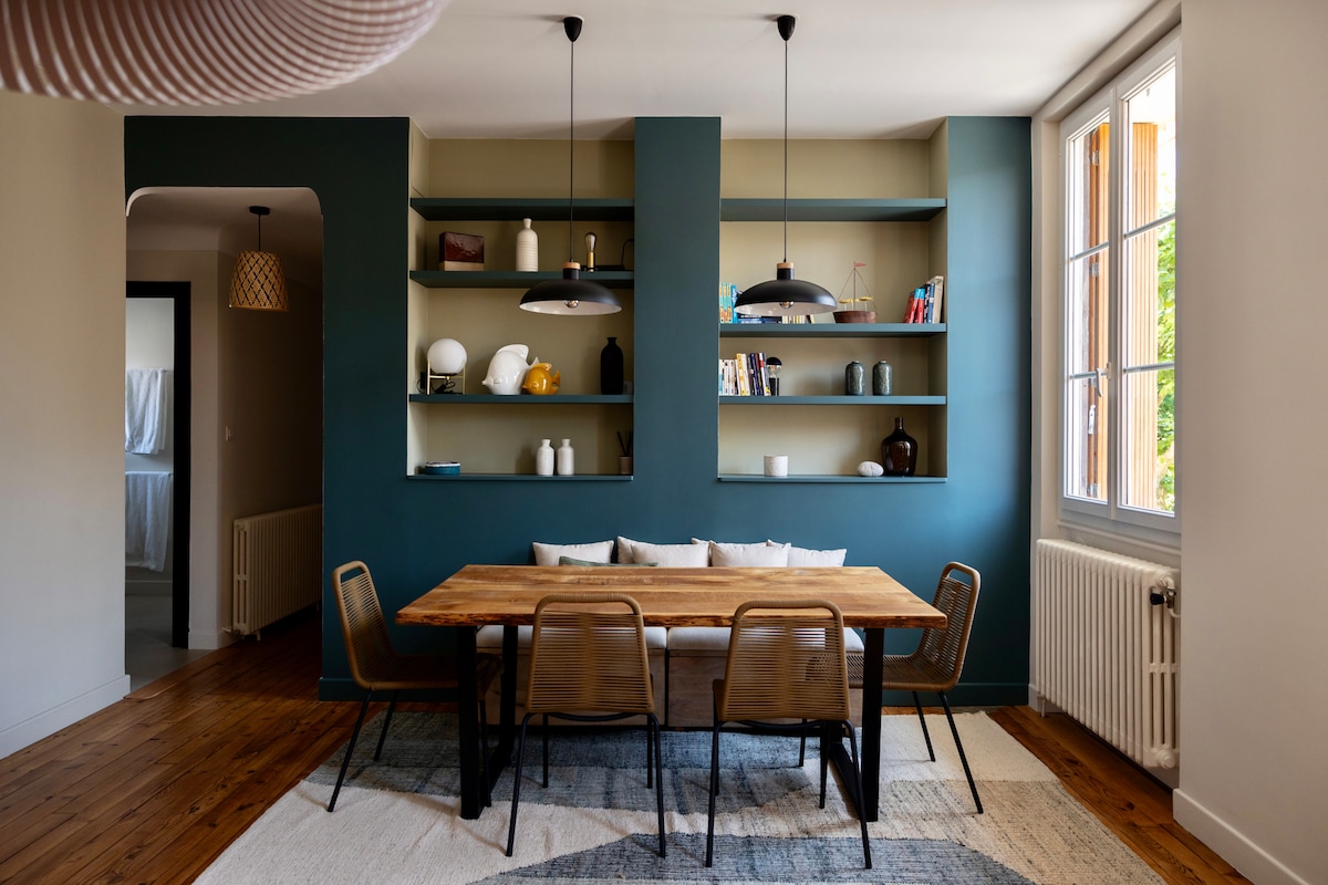 A spacious dining area is showcased with a wooden table surrounded by four metal chairs. The feature wall is painted in a deep blue, highlighting several shelves displaying decorative objects. Large windows allow natural light to fill the room, enhancing its open atmosphere.