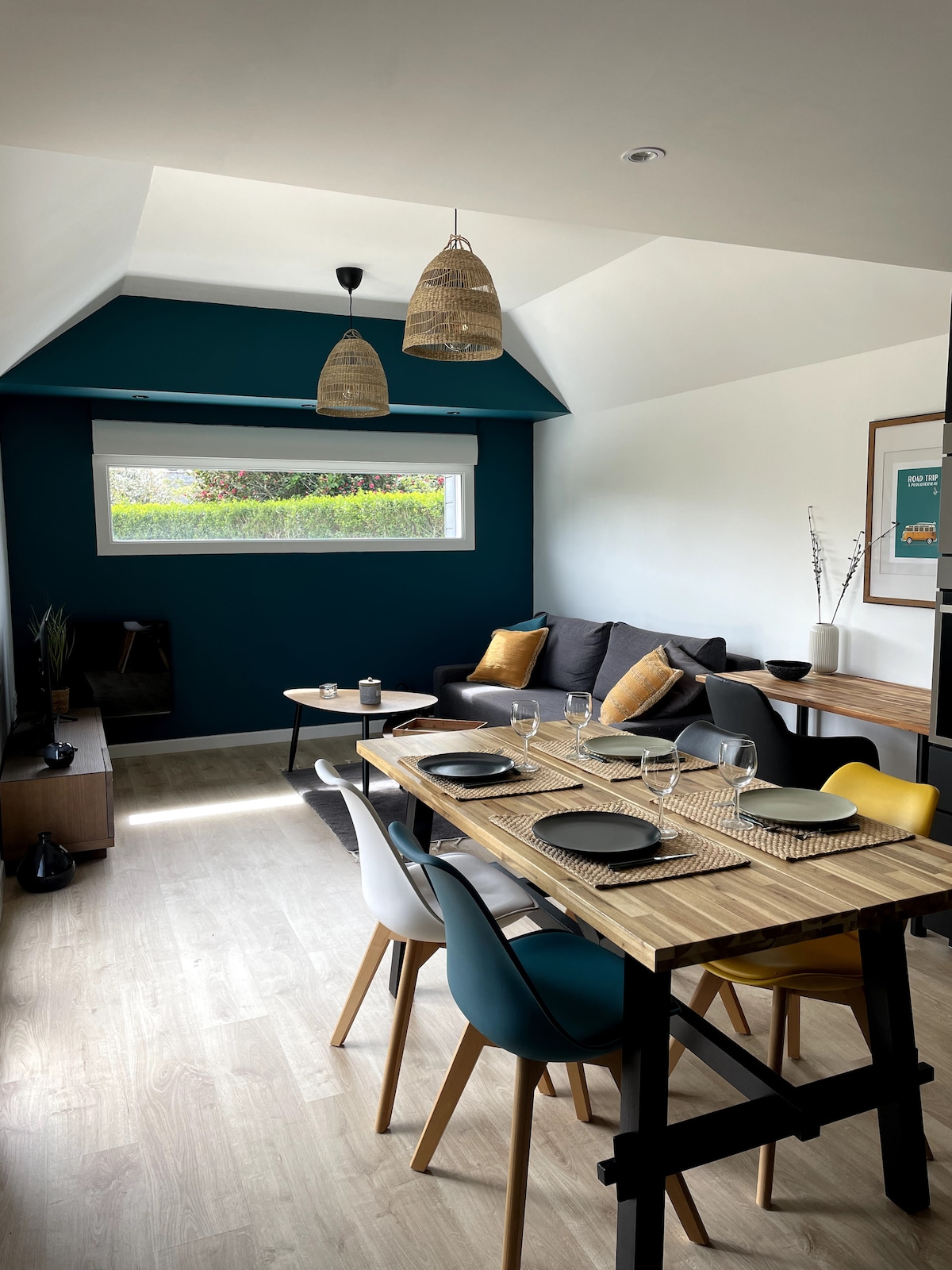 A modern living and dining area is presented with a high ceiling and large window offering natural light. A dining table with colorful chairs is set for four. A comfortable sofa is arranged nearby, paired with decorative cushions. Two woven pendant lights hang above the space.
