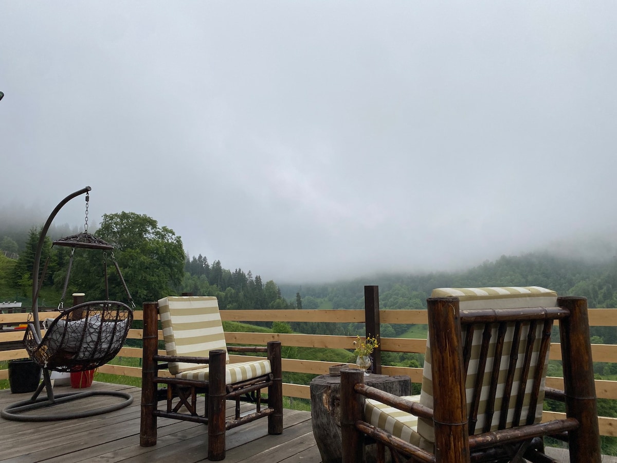 A spacious terrace features comfortable wooden furniture, including striped chairs and a hanging chair. The railing provides a view of the lush, fog-covered mountains and greenery beyond, creating a serene outdoor setting. A rustic table with a small flower adds a touch of nature.
