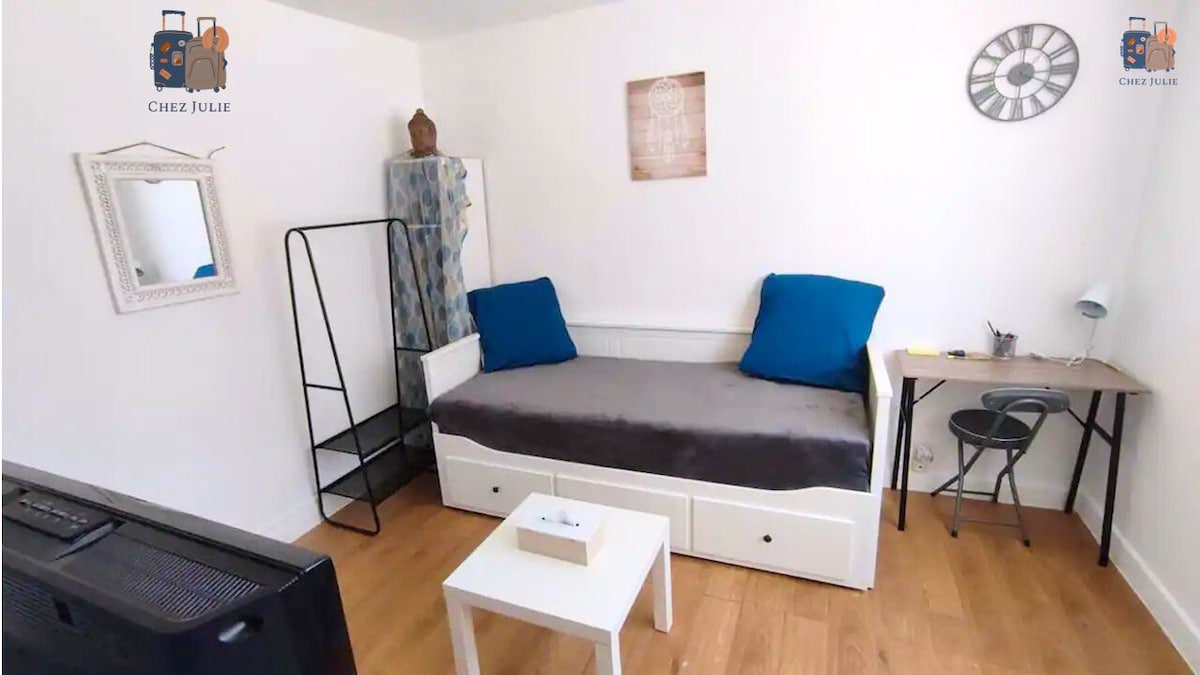 A bright, minimalist living space features a single bed layered with a soft gray blanket and accented by blue cushions. A small desk with a chair and a shelving unit are present, alongside a wall clock and a decorative mirror, highlighting a functional design.