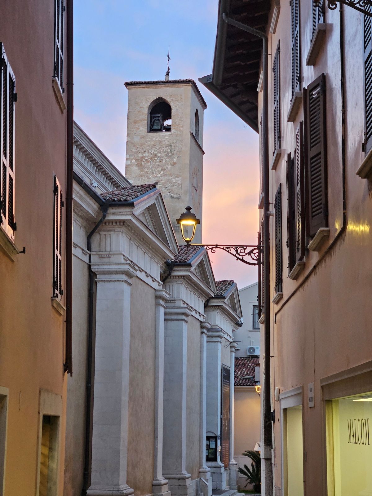 A narrow alleyway is framed by historic buildings, revealing a bell tower in the distance. The warm glow of a street lamp adds to the ambiance as pastel colors fill the sky at sunset, highlighting the charming architecture.