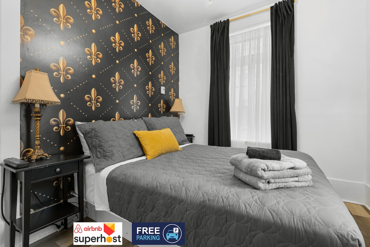 A cozy bedroom showcases a spacious bed with a grey quilt and layered towels at its foot. The backdrop features a dramatic patterned wall in black and gold, complemented by nightstands with elegant lamps. Curtains frame a window, allowing natural light to enter the room.