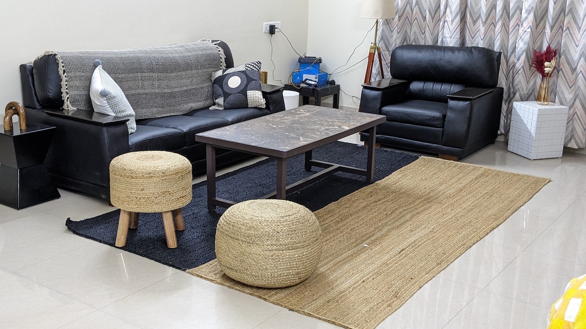 The living area is furnished with a comfortable black sofa and an accent chair. A rectangular coffee table sits atop a natural fiber rug, surrounded by woven ottomans. Curtains with a geometric pattern frame the window, and a lamp adds soft lighting to the space.