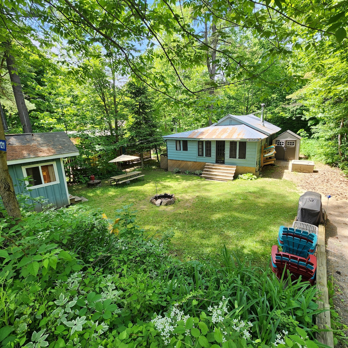 A peaceful outdoor setting features a cottage surrounded by lush greenery. A firepit sits in the foreground, with seating areas visible nearby. The cottage features a rustic façade with steps leading to its entrance. A small shed is also present, enhancing the natural ambiance.