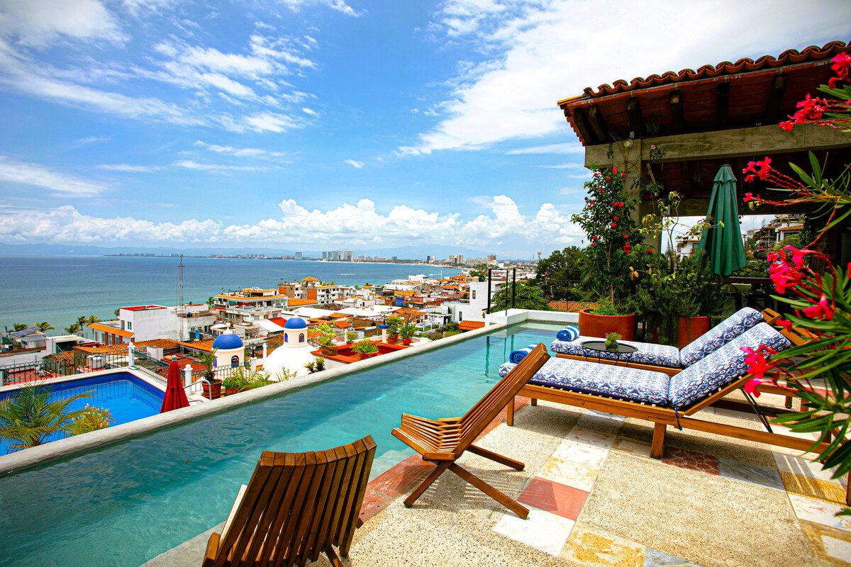The rooftop terrace features an infinity pool with expansive views of the ocean and city below. Lounge chairs are positioned alongside the pool, surrounded by vibrant plants and a shaded umbrella, creating a relaxing outdoor space for guests.