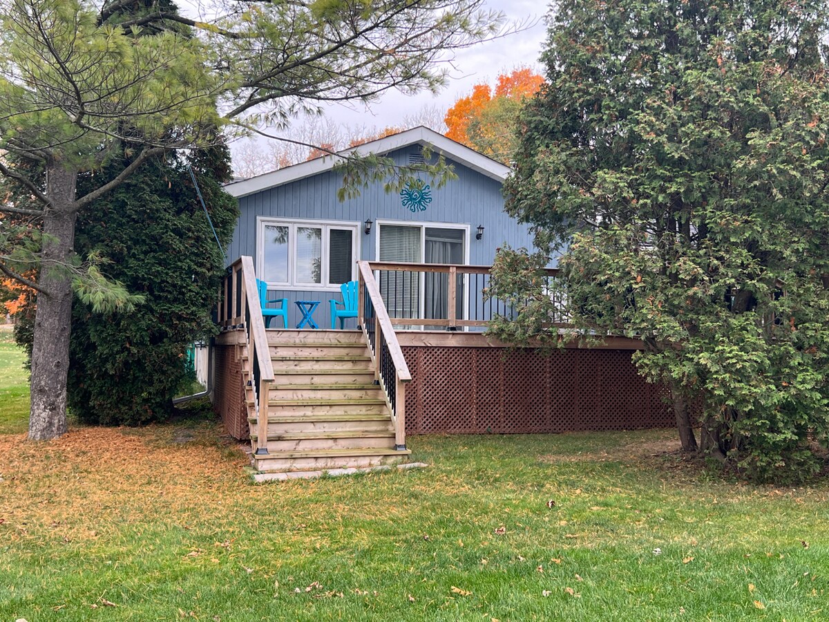 The exterior of a charming house is shown, featuring a wooden deck with light blue seating and steps leading up from the lush green lawn. Surrounding trees provide natural privacy, while hints of fall colors are visible in the background.
