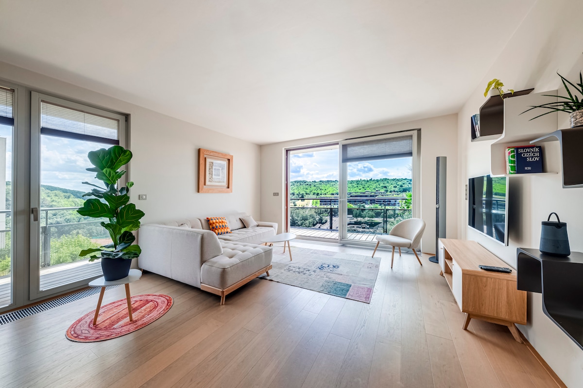 A bright living area features large glass doors opening to a scenic terrace. A comfortable sofa is paired with a stylish chair on light hardwood floors, while a decorative plant adds a touch of greenery. A wooden console table holds a television, completing the space.