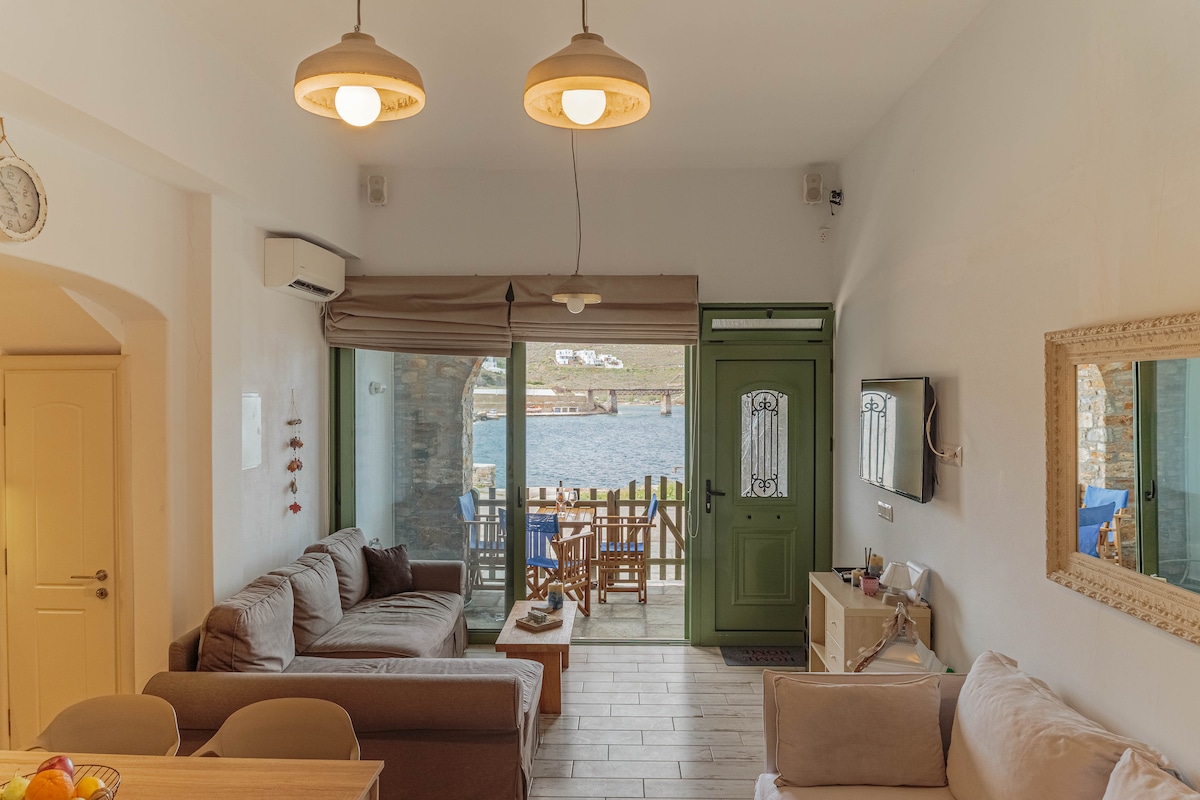 A bright living area features a comfortable sofa and a dining table for four. Large glass doors open to a balcony with sea views, while natural light fills the space. The decor includes subtle coastal elements, creating a relaxed ambiance.