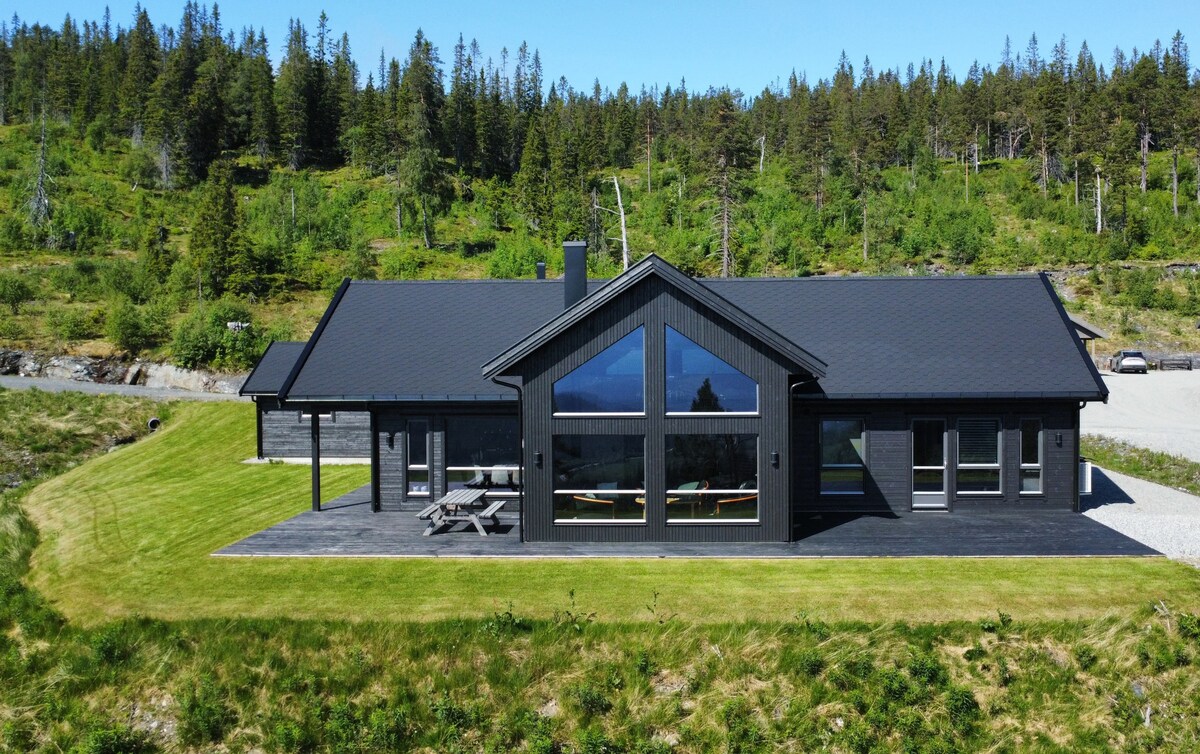The modern two-story cabin features a black exterior with large windows, allowing natural light to flood the interior. A spacious deck is visible, accompanied by picnic seating. Surrounded by greenery, the property is set in a tranquil area amidst trees and a grassy landscape.