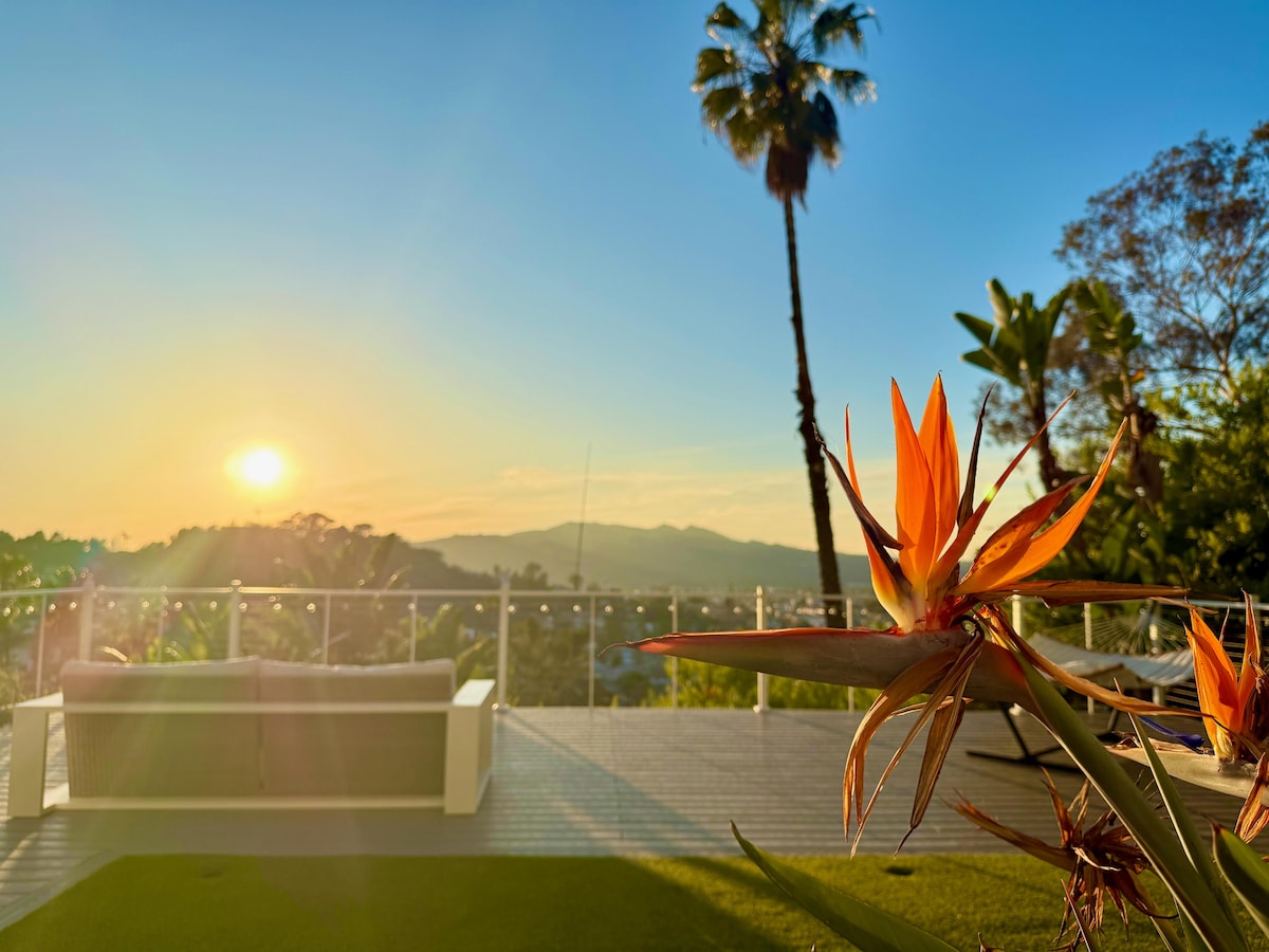 A vibrant bird of paradise flower is prominently displayed in the foreground, with a spacious deck visible behind it. The sun sets over distant hills, casting a warm glow across the landscape, while a sofa sits invitingly on the deck, offering a perfect spot for relaxation.