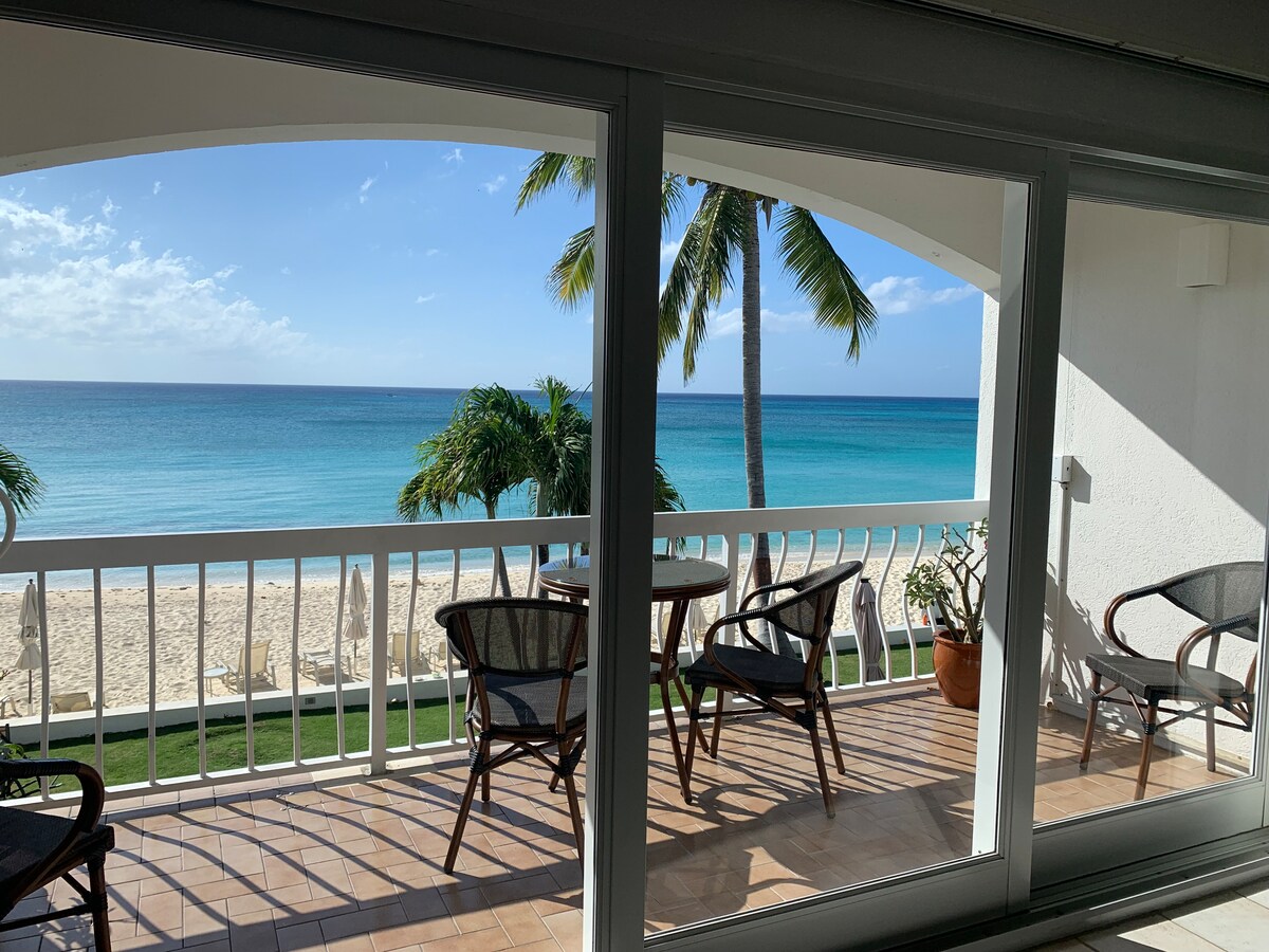 A balcony view showcases a tranquil Caribbean Sea, with soft waves gently lapping the shore. Palm trees frame the scene, while a set of four wicker chairs and a small table sit on the patio, offering a relaxed space to enjoy the beachside ambiance.