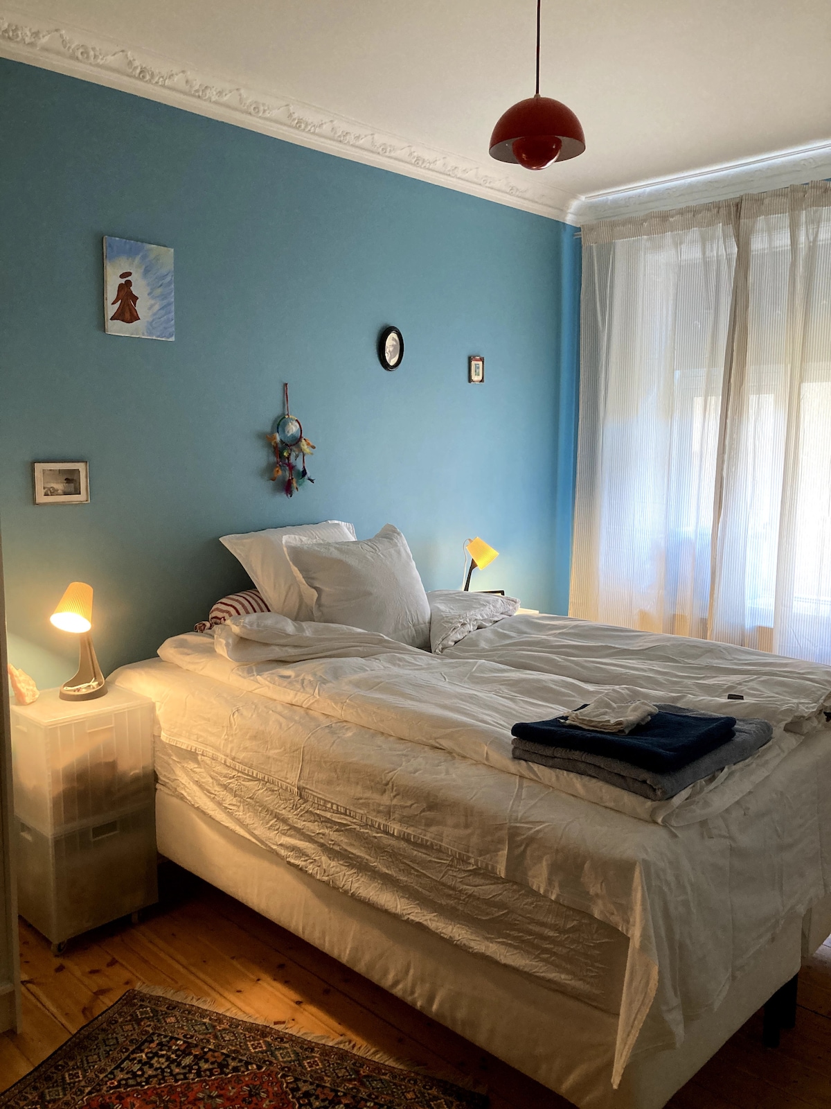A calming bedroom features a spacious bed with white bedding, set against a soft blue wall. Gentle light from bedside lamps and a window with sheer curtains brightens the room. Decorative elements include small framed pictures and a hanging dreamcatcher.